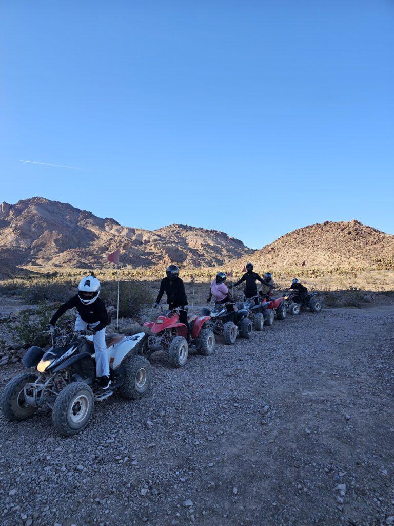 Six riders on red and white ATVs explore a rocky Nevada desert trail near Las Vegas with mountains and blue sky in the background.