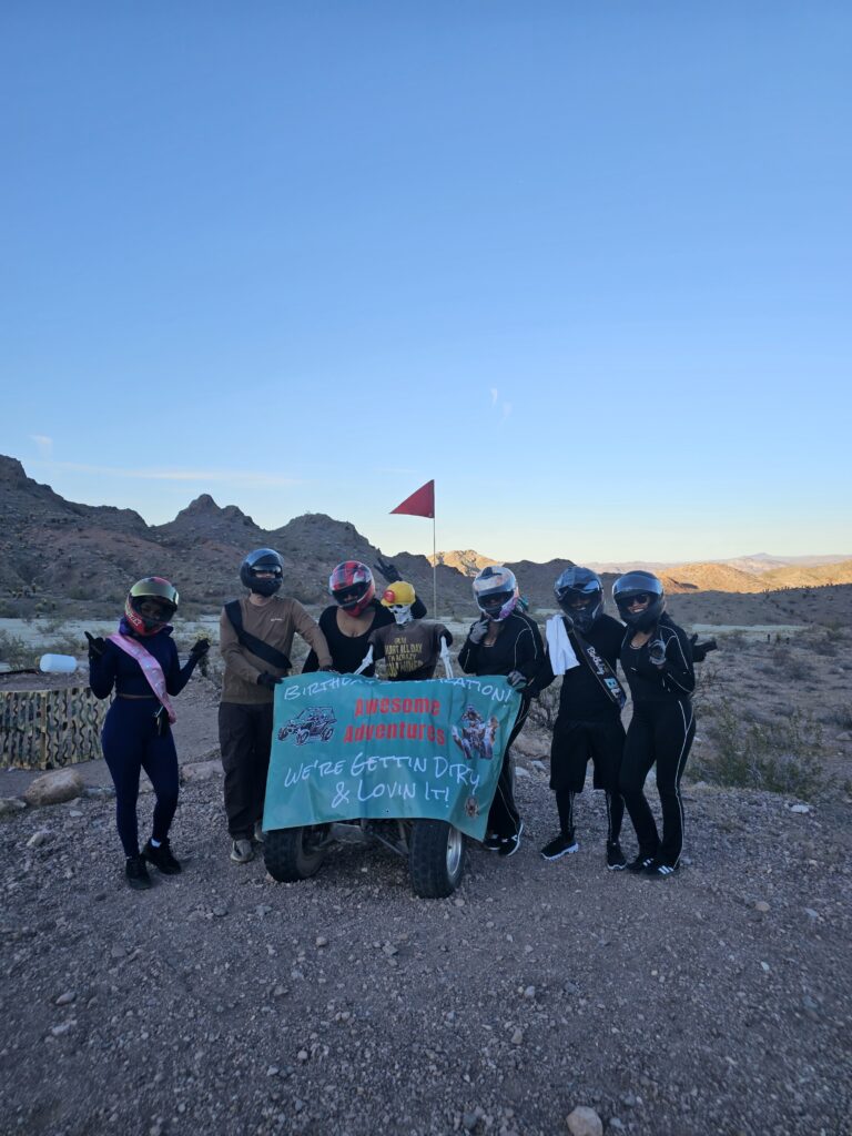 Group in helmets pose by ATV with “Bachelorette Adventure” banner near Las Vegas on a Nevada desert RZR off-road tour.