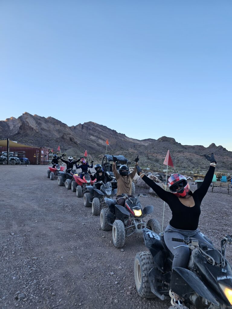 Adventurers on ATV tours ride in a line through Nevada desert trails near Las Vegas, smiling with mountains in the background.