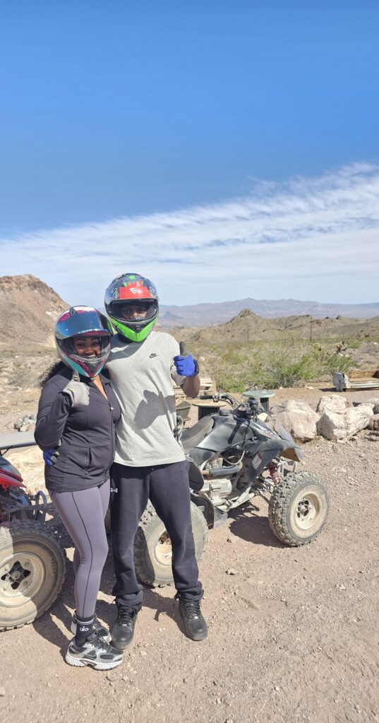 Smiling pair in helmets pose by ATVs on a Nevada desert trail near mountains—Las Vegas ATV tour adventure.