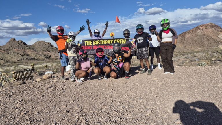 Adventurers in helmets pose with a "The Birthday Crew" banner during a Nevada ATV tour near Las Vegas, rocky desert and sky behind.