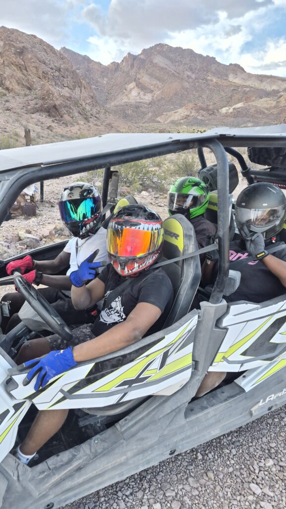 Group in helmets enjoys a Nevada ATV tour near Las Vegas, riding an RZR on rocky trails with mountains and a peace-signing driver.