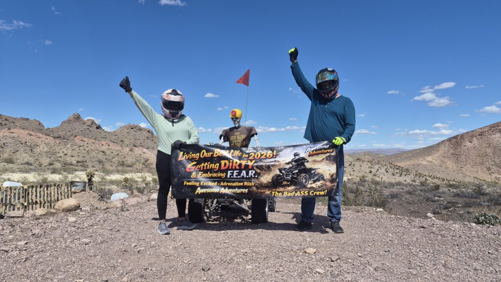 Adventurers in helmets celebrate atop a Nevada hill, banner raised, RZR behind them; Las Vegas ATV tour with desert and mountains views.