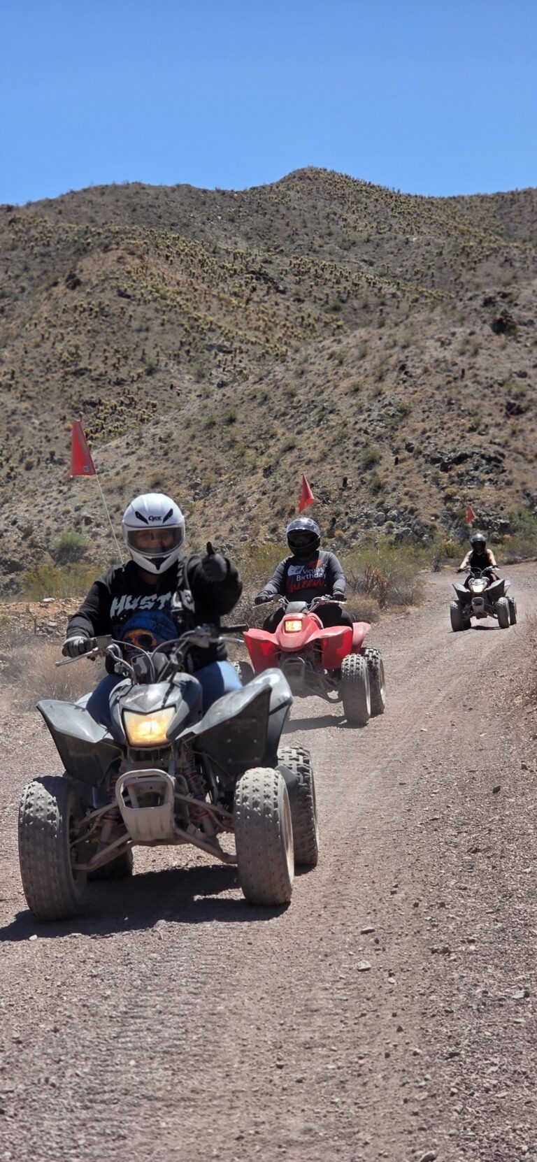 ATV riders with red flags explore a dusty Nevada desert trail near Las Vegas, mountains in view—perfect for guided ATV tours.