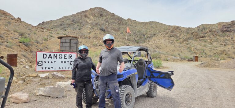 Adventurers in helmets by a blue ATV near Las Vegas, Nevada, explore a rocky trail with warning sign on an RZR off-road tour.
