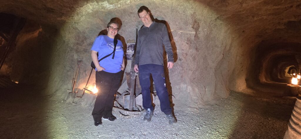 Two people smile in a Nevada underground tunnel near mining gear—perfect for Las Vegas ghost town sightseeing and ATV adventures.