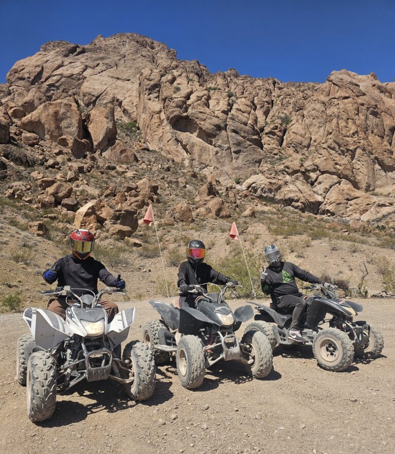 Riders in helmets on ATVs with red flags explore a Nevada desert trail near rocky cliffs under blue skies, ideal for Las Vegas ATV tours.