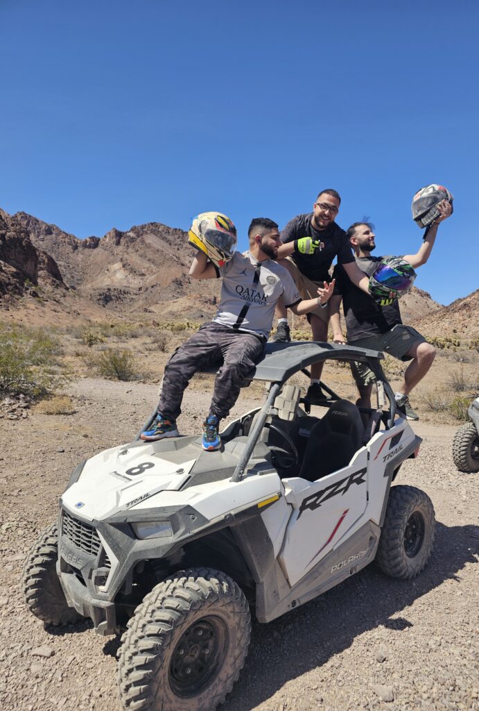 Three men with helmets pose by a Polaris RZR in the Nevada desert near Las Vegas on an ATV tour under clear blue skies.