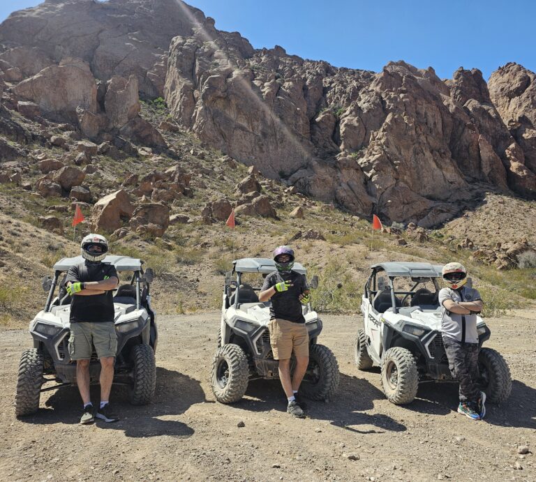 Adventurers in helmets pose by three ATVs in Nevada desert near Las Vegas mountains, ready for a RZR off-road tour.