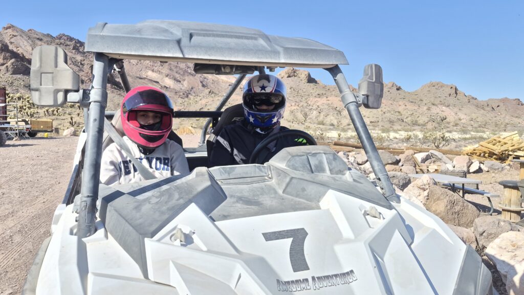Two people in helmets ride a white RZR ATV with #7, exploring Nevada desert near Las Vegas on an off-road adventure tour.
