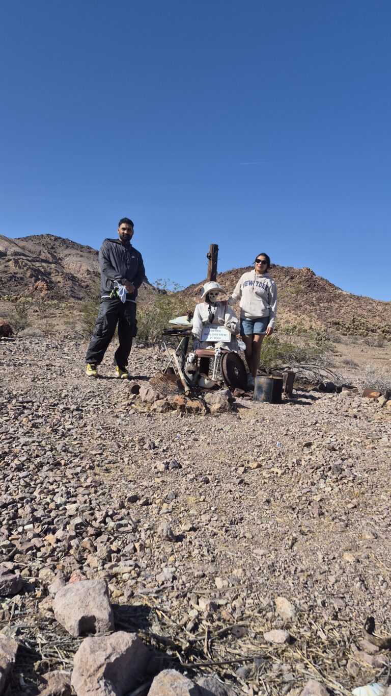 Two people explore a Nevada ghost town near Las Vegas, beside a metal skeleton sculpture and rusty relics, mountains in the background.