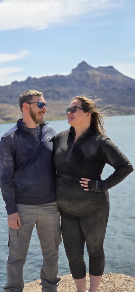 Couple in sunglasses on rocky Colorado River shore near Las Vegas, Nevada, smiling with mountains—perfect for ATV tours and sightseeing.