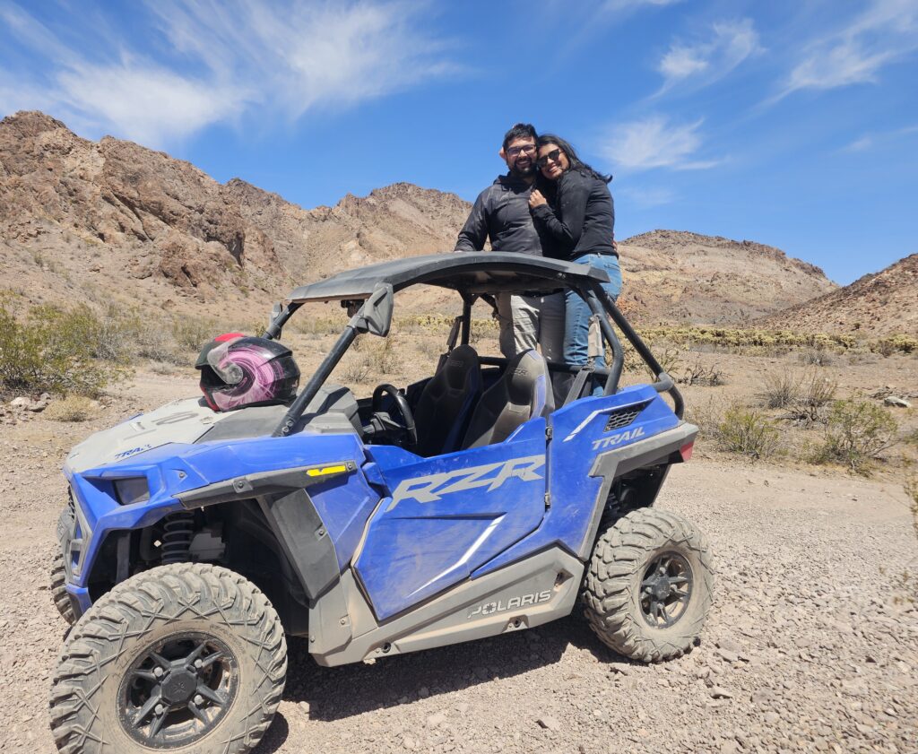 Smiling friends hug atop a Polaris RZR during a Nevada ATV tour near Las Vegas; pink helmet on seat, desert views behind them.