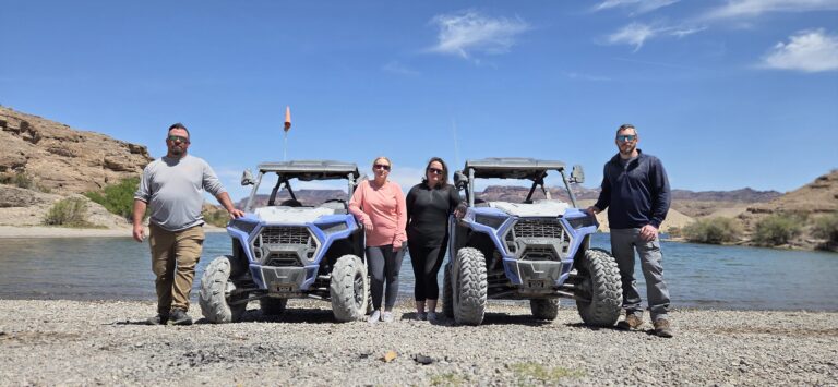 Group enjoys ATV adventure on Colorado River near Las Vegas, Nevada; RZR vehicles on rocky bank with scenic desert hills behind.
