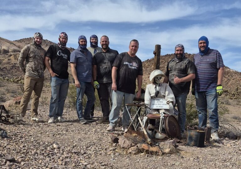 Eight men pose by a miner mannequin in Nevada desert near Las Vegas, ready for ATV tours with mountains and clouds behind.