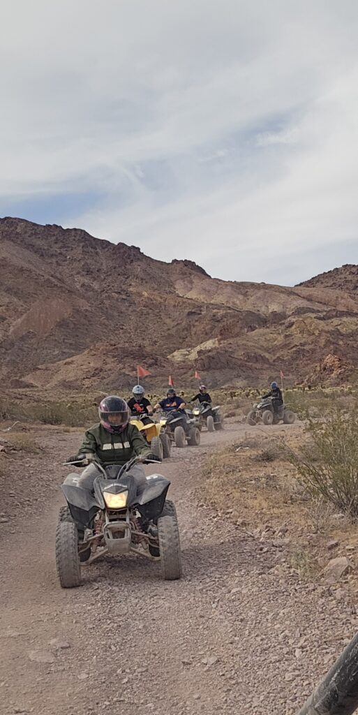 ATV riders with helmets explore a dusty Nevada desert trail near Las Vegas, surrounded by mountains and sparse vegetation.