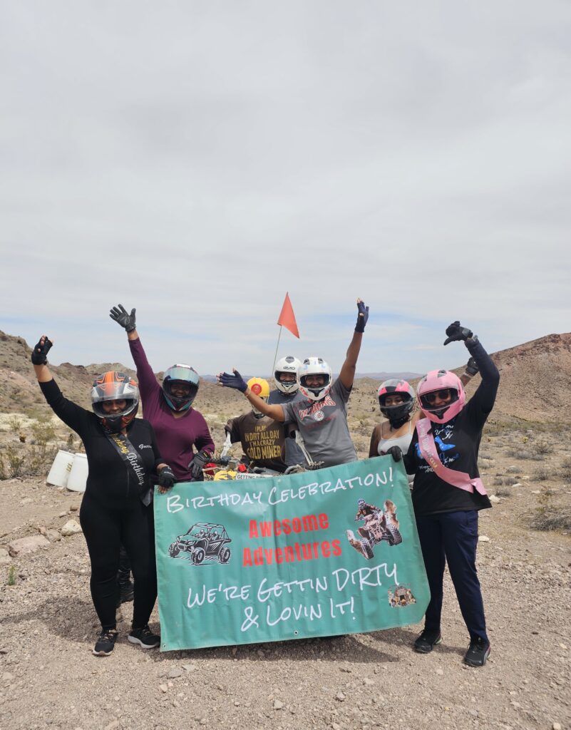 Group in helmets celebrates with a Birthday banner on a Nevada ATV tour near the Colorado River, desert landscape behind them.