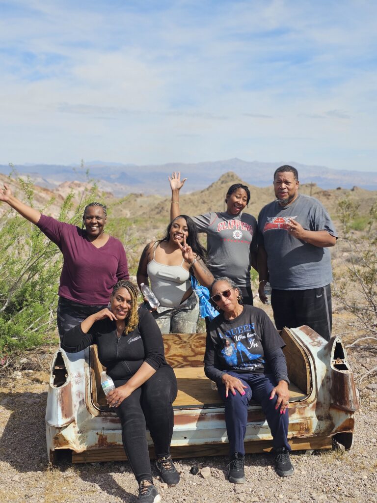 Five friends smile in the Nevada desert near Las Vegas, mountains behind, some on a rusty car seat—perfect for ghost town adventures.
