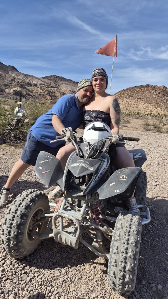 Smiling couple on an ATV in Nevada desert near Las Vegas, ready for RZR off-road tours with rocky hills and blue sky backdrop.