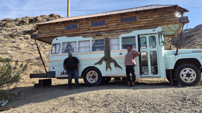Two people by a tree-mural bus cabin in Nevada desert near Las Vegas, with rocky hills—perfect for ATV tours or Ghost Town sightseeing.