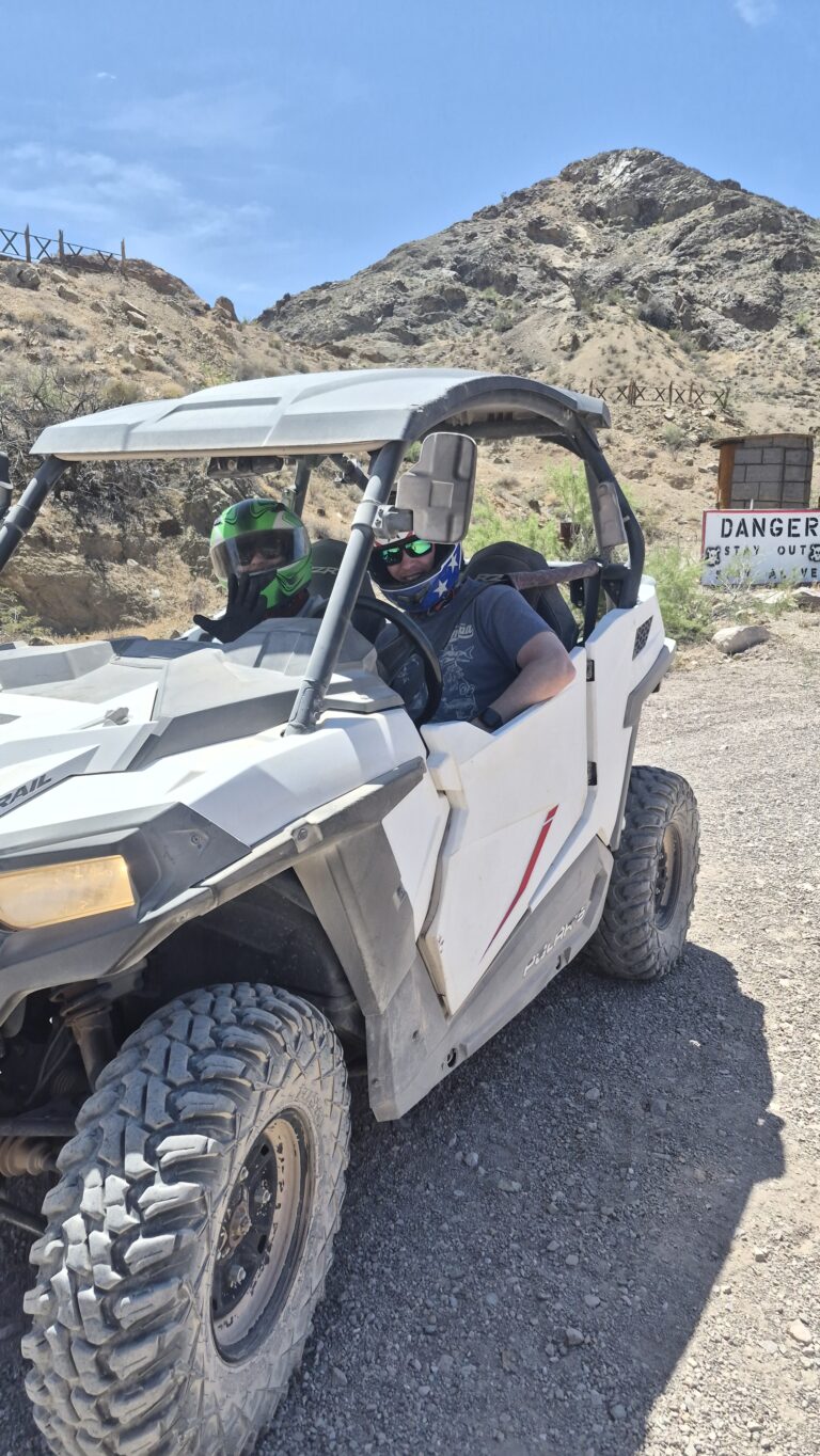 Riders in helmets enjoy a Nevada RZR off-road tour near Las Vegas, driving rocky trails past hills and a Danger sign under blue skies.
