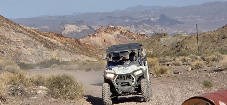 ATV tour near Las Vegas: white off-road vehicle on a dusty Nevada trail, riders in helmets, desert mountains in the background.