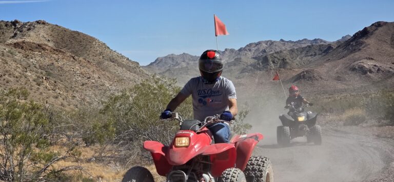 Riders on red ATVs explore a dusty Nevada trail near Las Vegas with helmets, safety flags, and mountain views in the background.