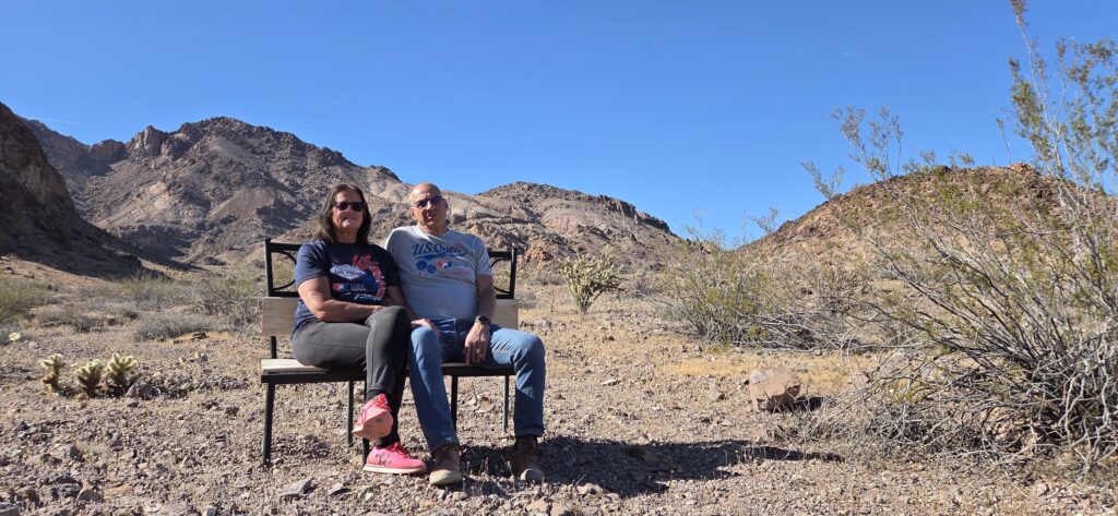 A couple relaxes on a bench in Nevada’s rugged desert near Las Vegas, with distant mountains and sparse vegetation under clear skies.