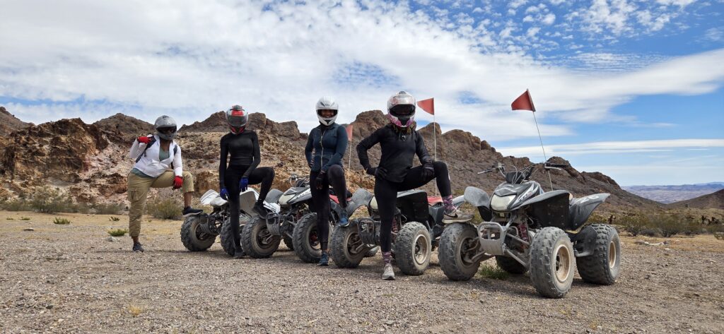 Group in helmets poses with ATVs on rocky Nevada desert near Las Vegas; red flags flying, mountains behind. ATV tours adventure.