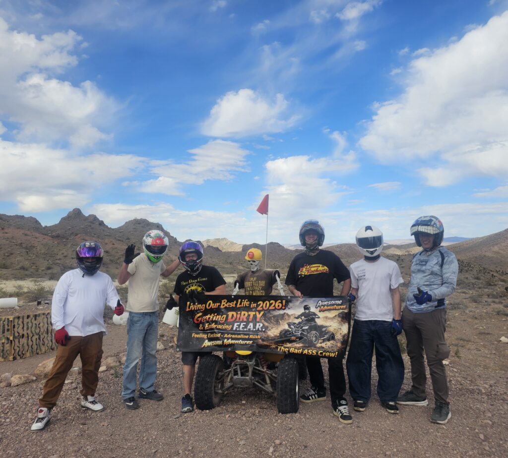 Group in helmets poses with ATV and adventure banner on rocky Nevada terrain; Las Vegas ATV tour, desert mountains behind.