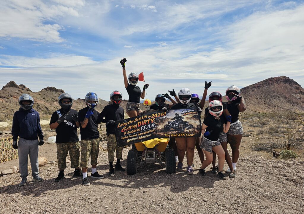 Adventurers on an ATV tour near Las Vegas pose with a banner in the Nevada desert, mountains and blue sky behind.