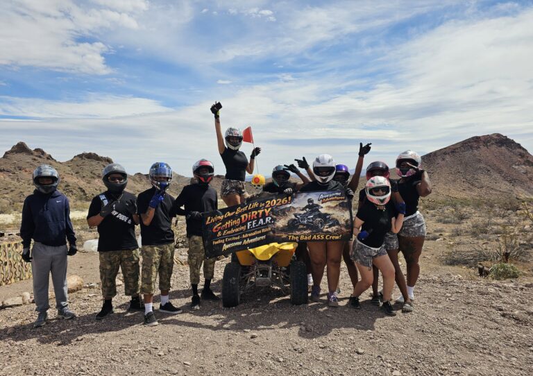 Adventurers on an ATV tour near Las Vegas pose with a banner in the Nevada desert, mountains and blue sky behind.