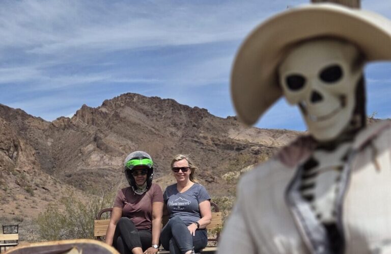 Skeleton in a hat with sign sits near rusty relics; two people bench-sit by Nevada desert mountains, perfect for ghost town tours.