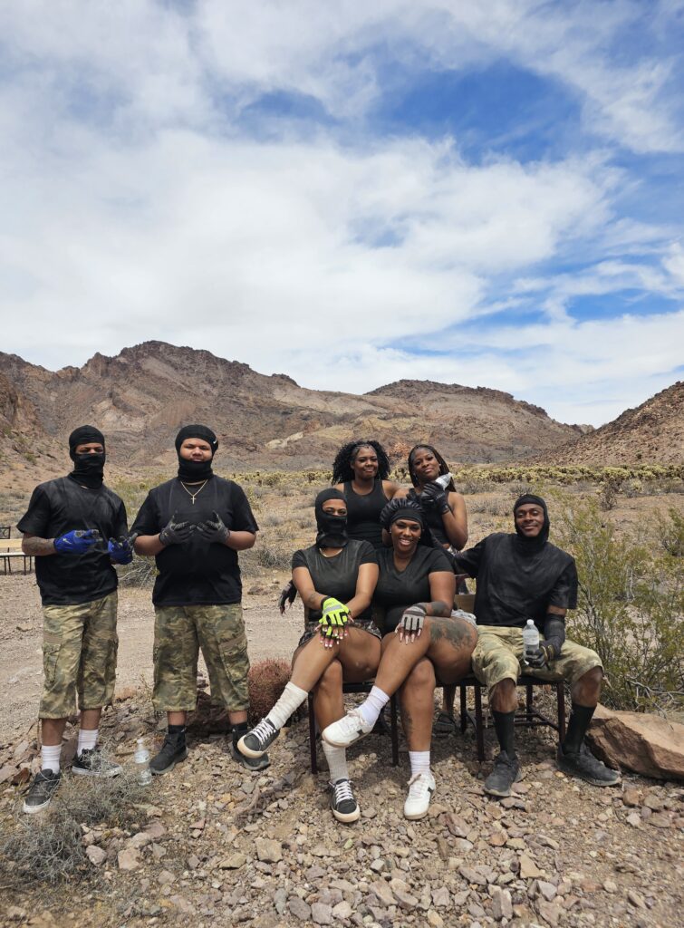 Group in black shirts and camo shorts pose in Nevada desert near Las Vegas before an ATV tour; mountains and rocky terrain behind.