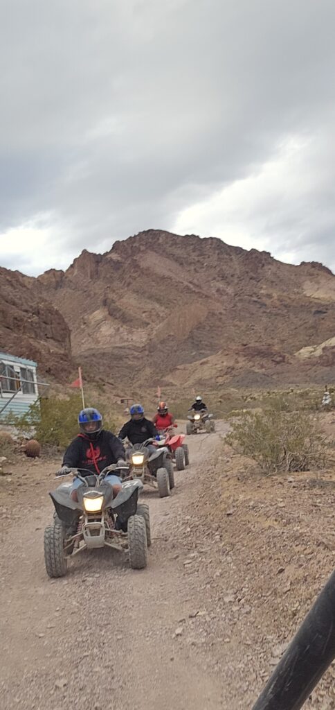 Four people on an ATV tour near Las Vegas ride along a desert trail with mountains, shrubs, and a building under cloudy Nevada skies.