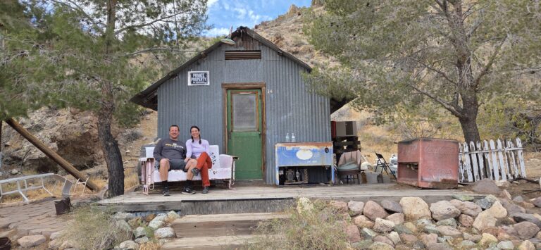 Two people relax on a bench by a rustic Nevada cabin, near desert trees and an old Pepsi cooler—perfect for Ghost Town sightseeing.