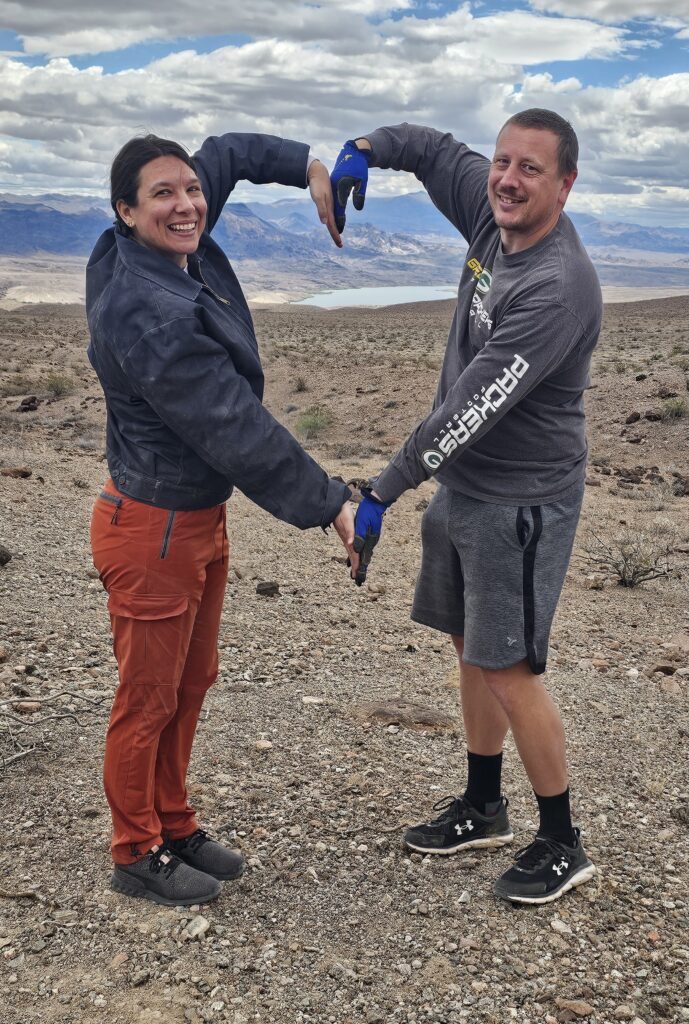 Smiling duo forms a heart on rocky Nevada terrain, with Las Vegas views and Colorado River in the background under partly cloudy skies.