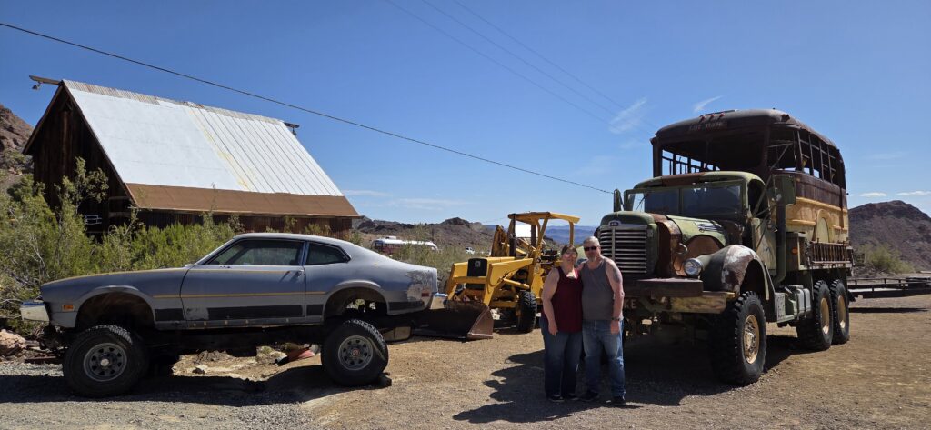 Two people in Nevada stand by a classic car and military truck, with desert sights near Las Vegas Ghost Towns in the background.