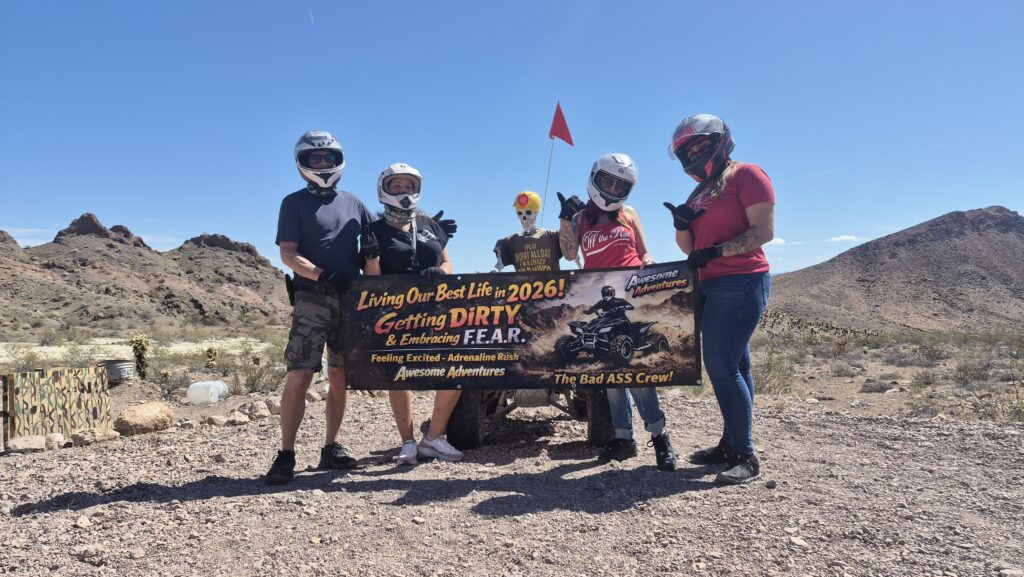 Group in helmets and casual wear holds banner in Nevada desert near Las Vegas, ready for ATV tours by Colorado River, mountains behind.