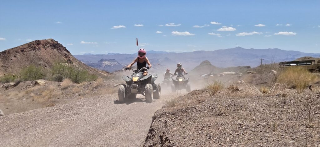 Riders on ATVs explore a dusty desert trail near Las Vegas, Nevada, with rocky hills and distant mountains under a blue sky.