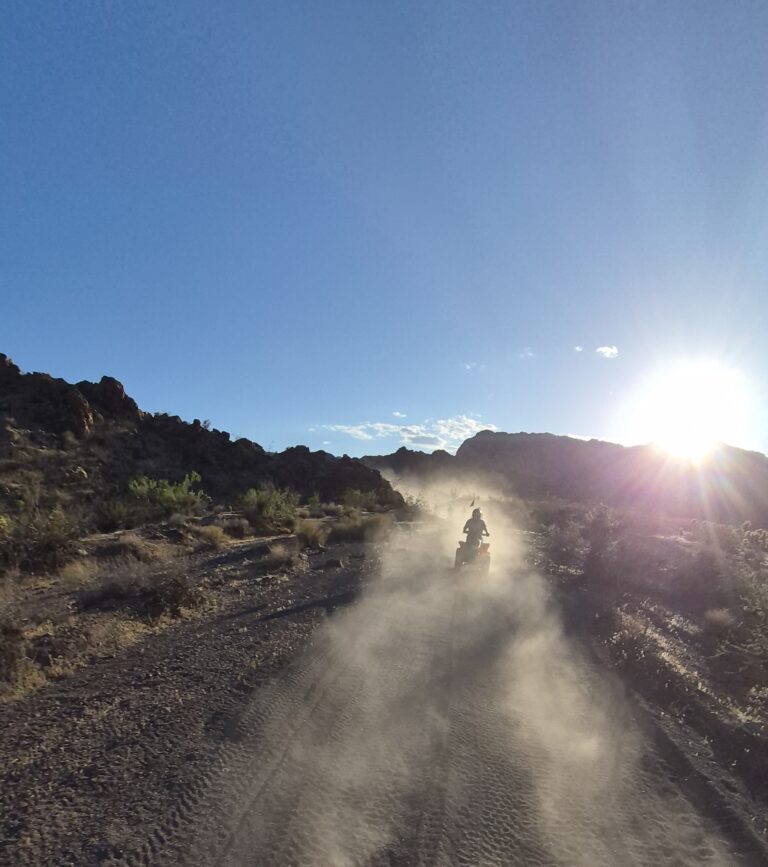 ATV rider kicks up dust on a Nevada desert trail at sunset near Las Vegas, with rocky terrain and desert shrubs in view.