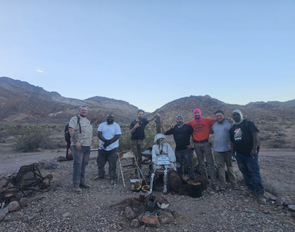 Seven people in Nevada pose with a dressed skeleton near mountains, enjoying a ghost town sightseeing tour by the Colorado River.