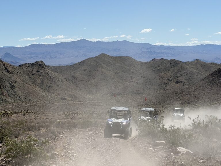 ATV tour group rides RZR off-road vehicles on a dusty Nevada desert trail near Las Vegas with mountains in the background.