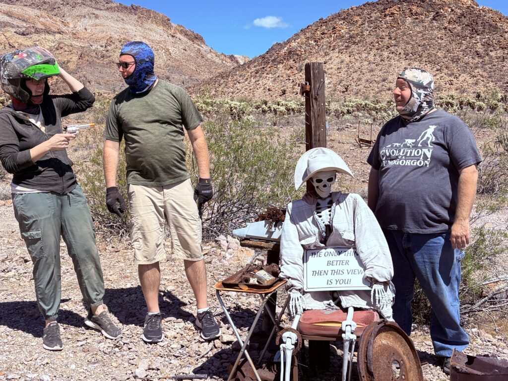 Adventurers in Nevada desert near Las Vegas pose by skeleton in wheelchair; mountains, blue sky & Ghost Town sightseeing vibes.