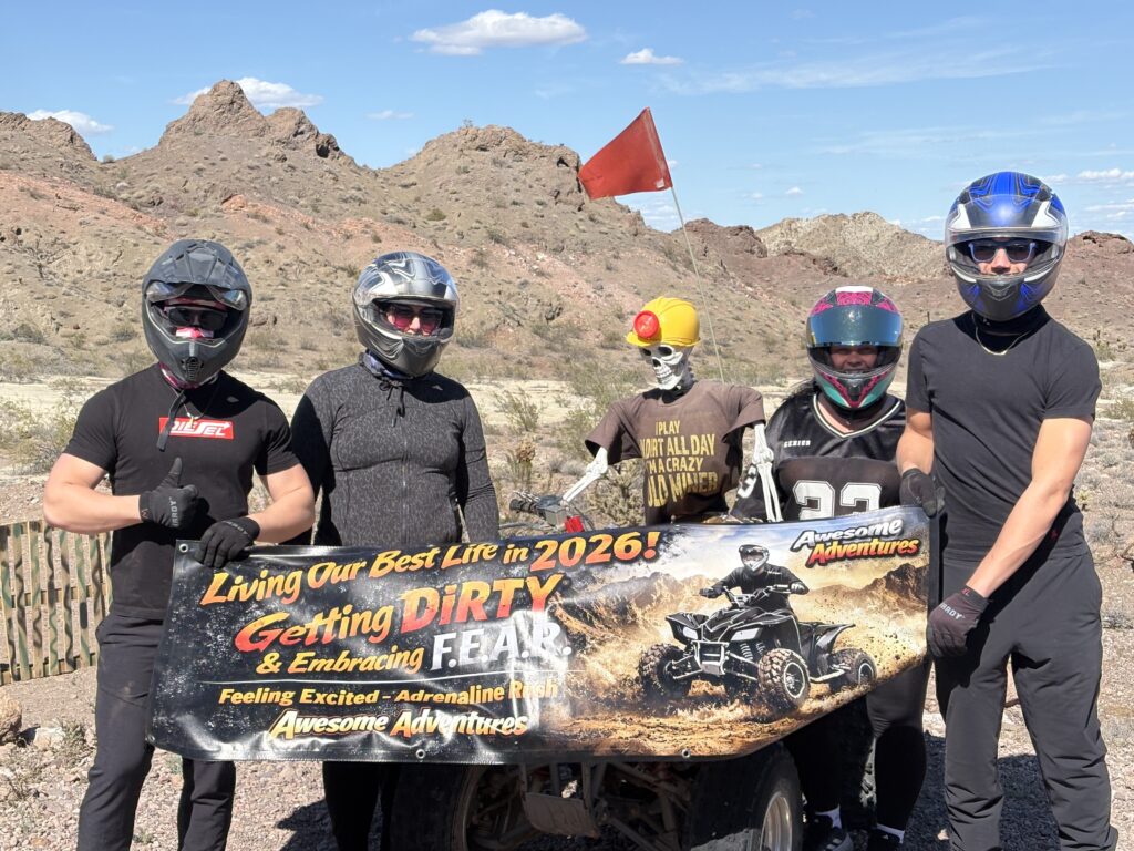 Group in helmets holds "Awesome Adventures" banner on Nevada desert ATV tour near Las Vegas; mannequin, red flag, rocky backdrop.