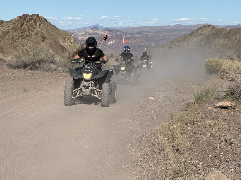 ATV riders in helmets speed along a dusty Nevada desert trail near Las Vegas on an exciting off-road RZR tour under blue skies.