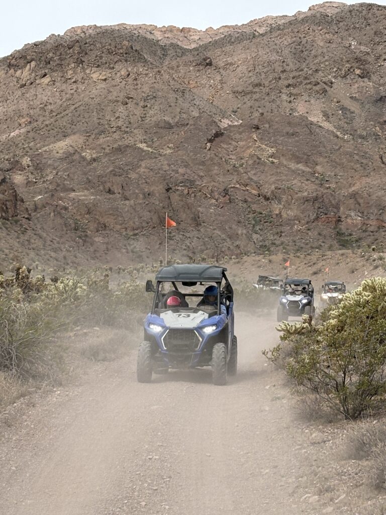 ATV and RZR off-road vehicles with orange flags drive near Las Vegas, Nevada, on a desert trail by rocky hills under cloudy skies.