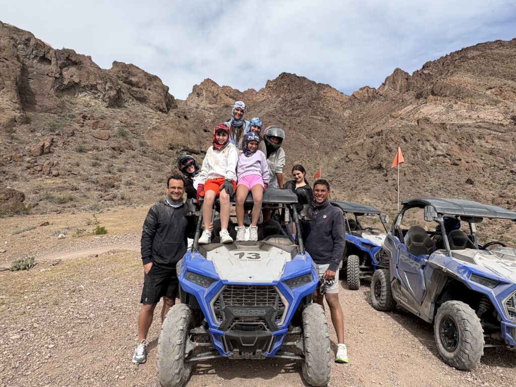 Adventurers pose on a blue RZR during a Nevada ATV tour near Las Vegas, with helmets, sunglasses, and desert mountains behind.