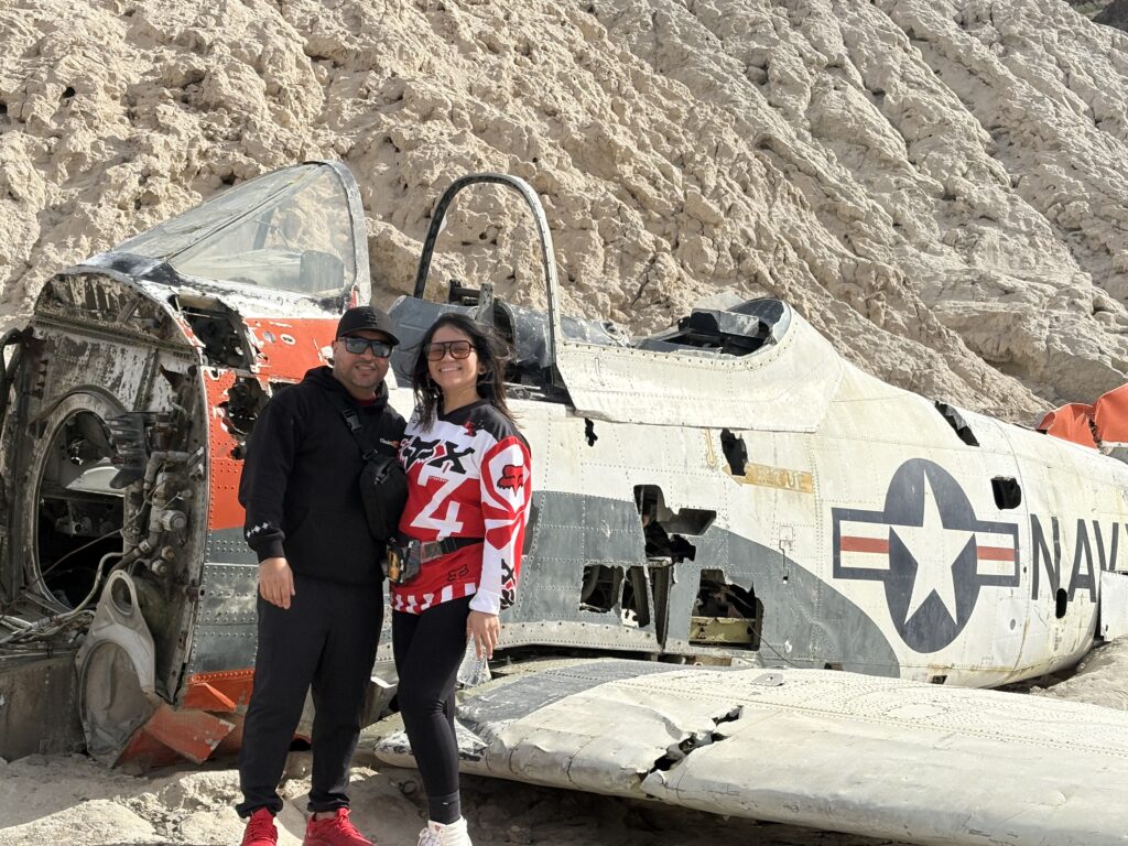 Smiling couple by a damaged Navy plane wreck in Nevada desert near Las Vegas Ghost Town, rocky hills in background.