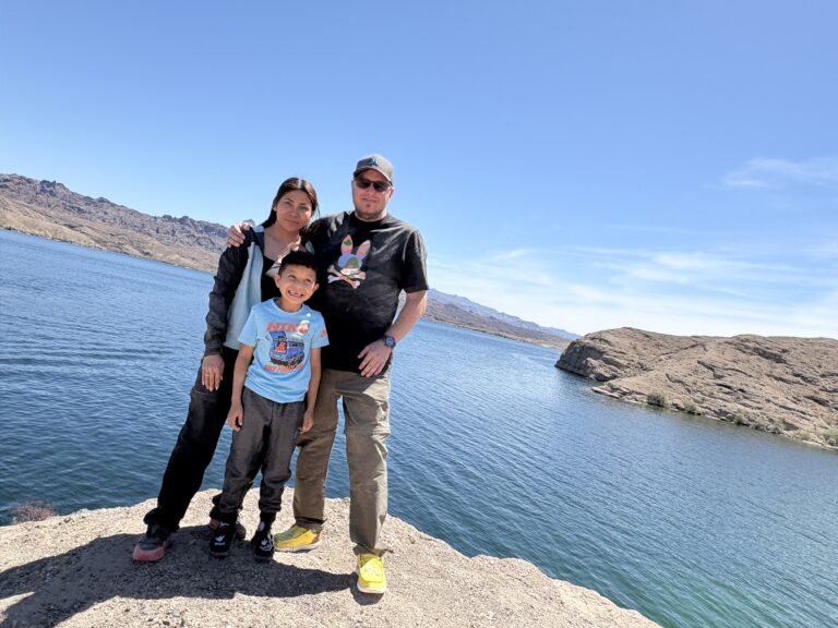 A family smiles on a rocky Nevada lakeshore near Las Vegas, with clear blue Colorado River water and scenic mountains behind them.